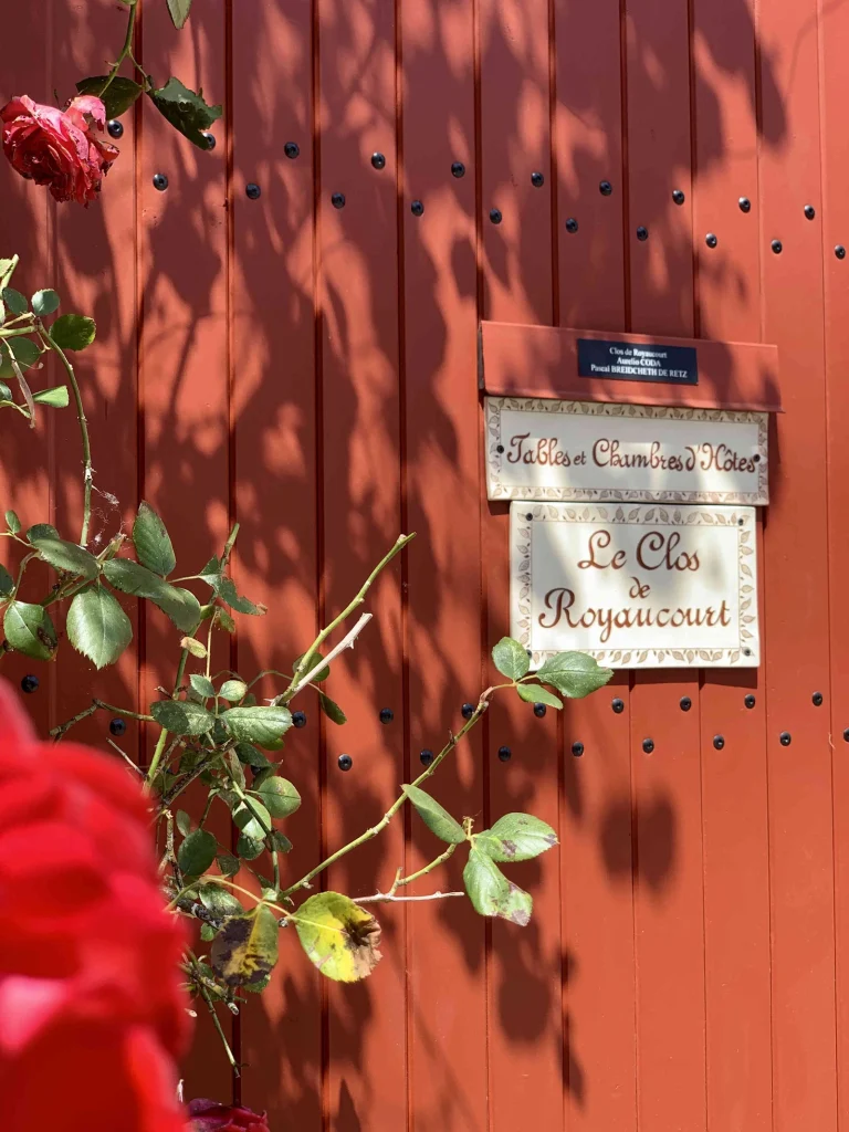 Portail en bois rouge à l'entrée du Clos de Royaucourt, avec ses enseignes « Chambres et Tables d'hôtes » et « Le Clos de Royaucourt » entourées de rosiers