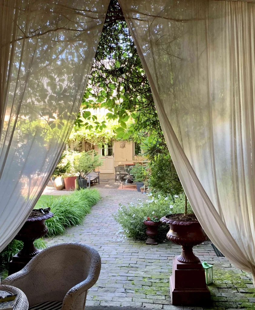 Vue depuis le porche du Clos de Royaucourt sur l'allée pavée du jardin, encadrée de voilages et de plantes en pot