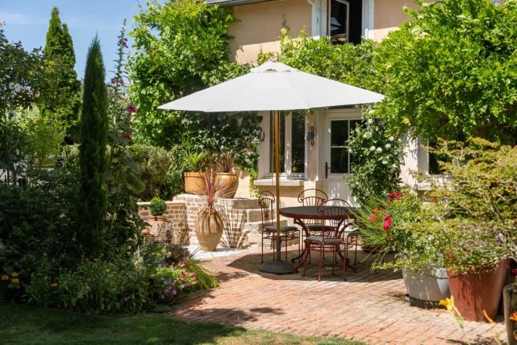 Terrasse ombragée du Clos de Royaucourt, salon de jardin sous un grand parasol blanc, façade en pierre picarde et rosiers grimpants